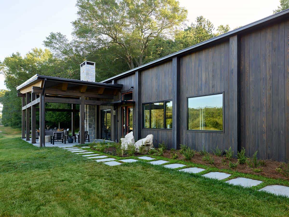 Side elevation of dark wood farmhouse with stone stepping path, covered porch, and large picture windows