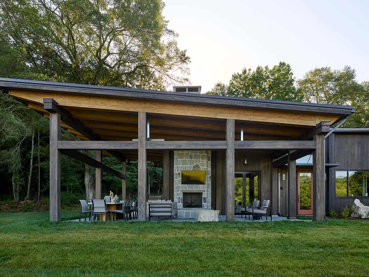 Covered outdoor living pavilion with exposed timber beams, stone fireplace, TV, and dining area on lawn