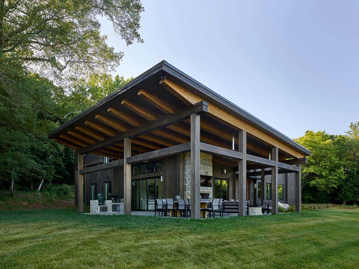 Rear view of timber-framed outdoor pavilion with exposed rafters, stone chimney, outdoor kitchen, and lawn