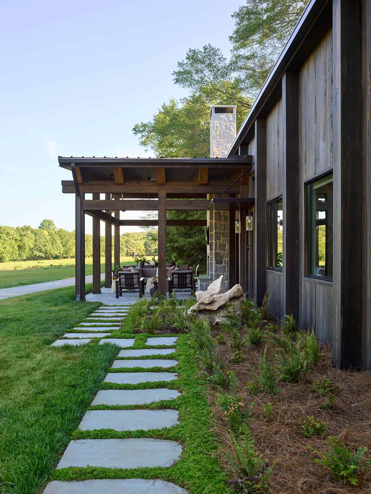 Flagstone stepping path leading to covered porch with timber pergola, stone chimney, and pastoral field views