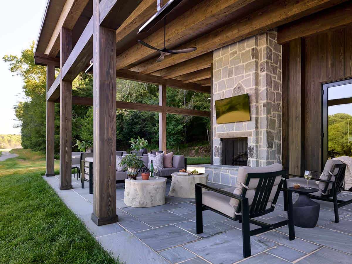 Covered outdoor room with timber columns, stone fireplace, black iron furniture, stump coffee tables, and ceiling fan