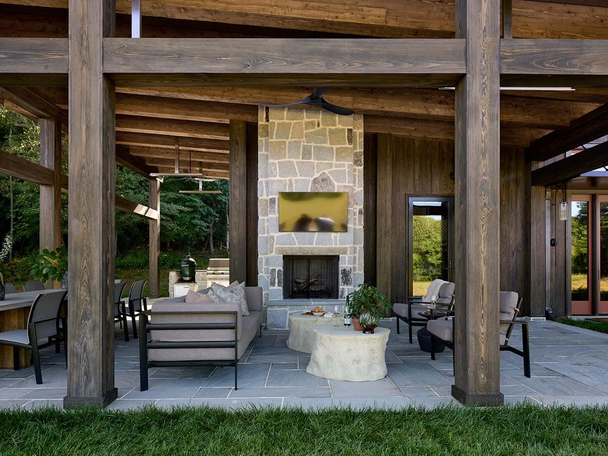 Open-air outdoor living area with timber beams, stone fireplace, grey sofas, outdoor kitchen, and red entry door