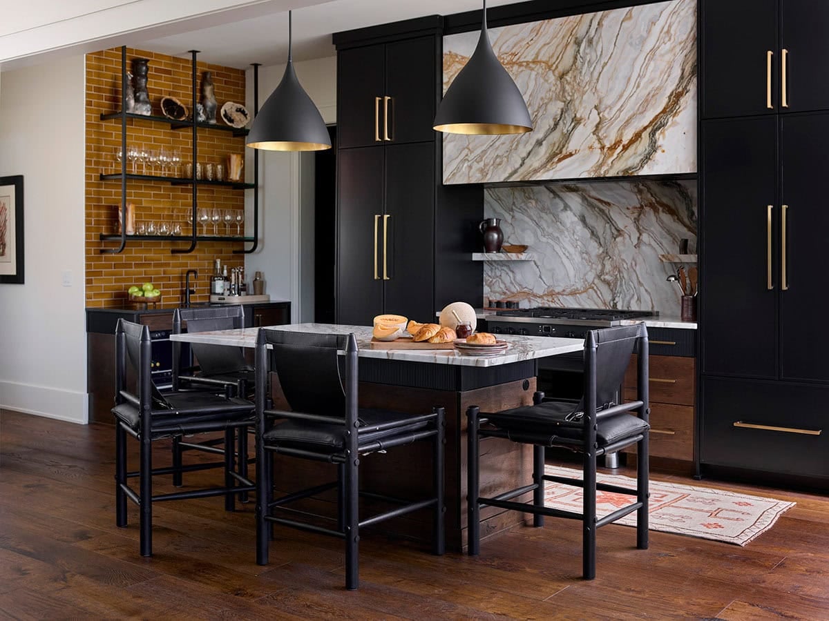 Dark contemporary kitchen with black cabinetry, brass hardware, veined marble backsplash, amber tile, and black pendant lights