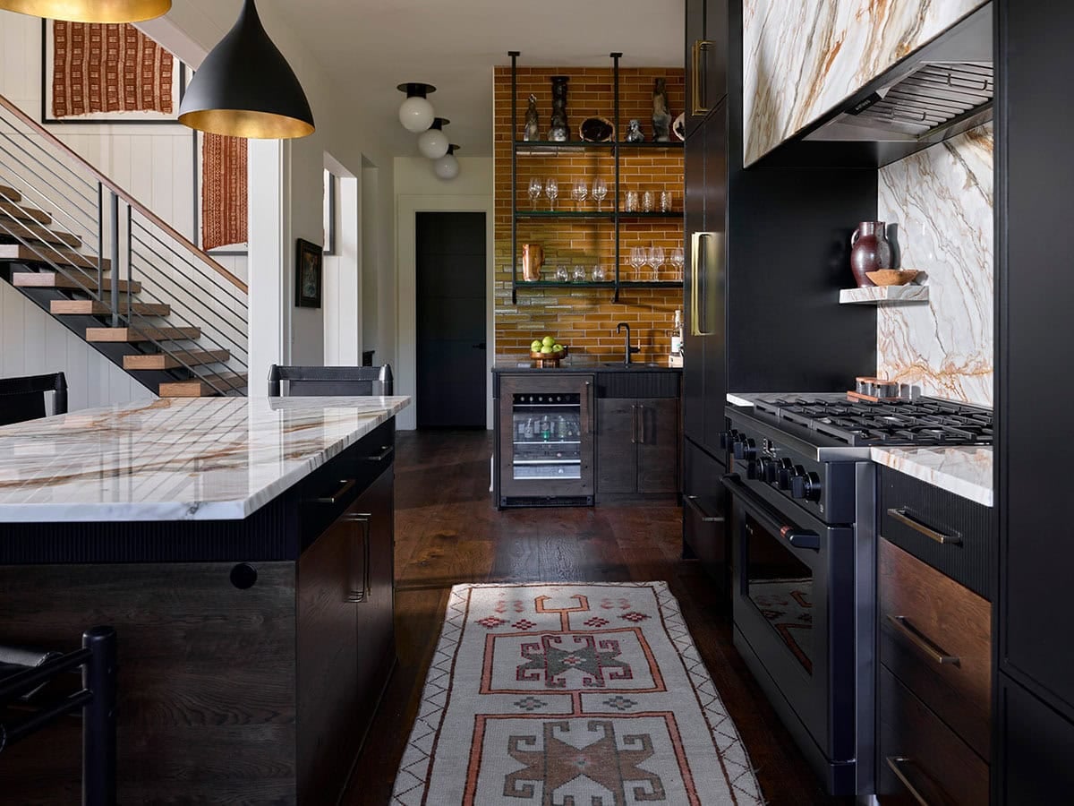 Kitchen and staircase view with marble island, professional range, amber tile bar area, vintage runner, and black metal stair railing