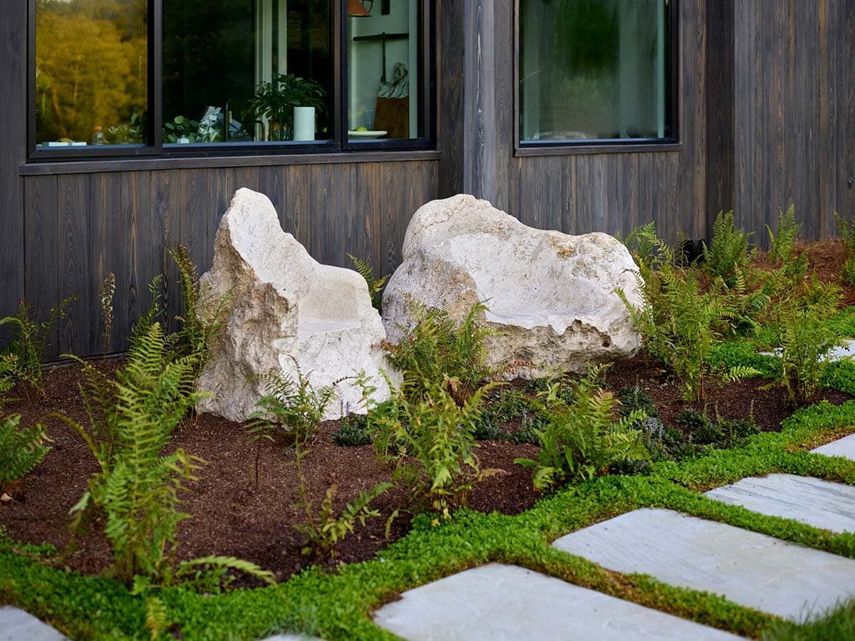 Large white limestone boulders nestled among ferns against dark wood exterior siding