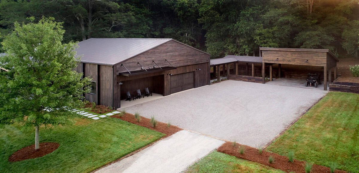 Aerial view of barn-style home with metal roof, gravel drive, carport, and wooded backdrop