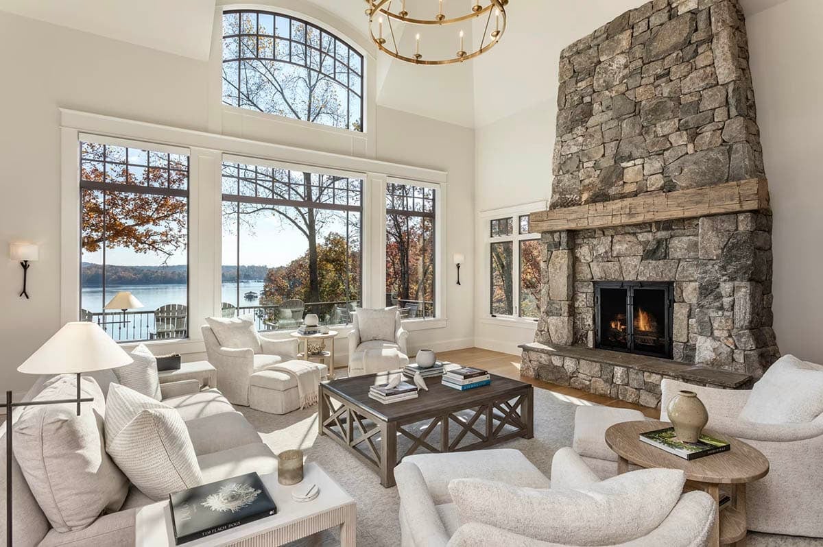 Bright lakeside living room with soaring ceilings, floor-to-ceiling windows, white sofas, and a floor-to-ceiling stone fireplace