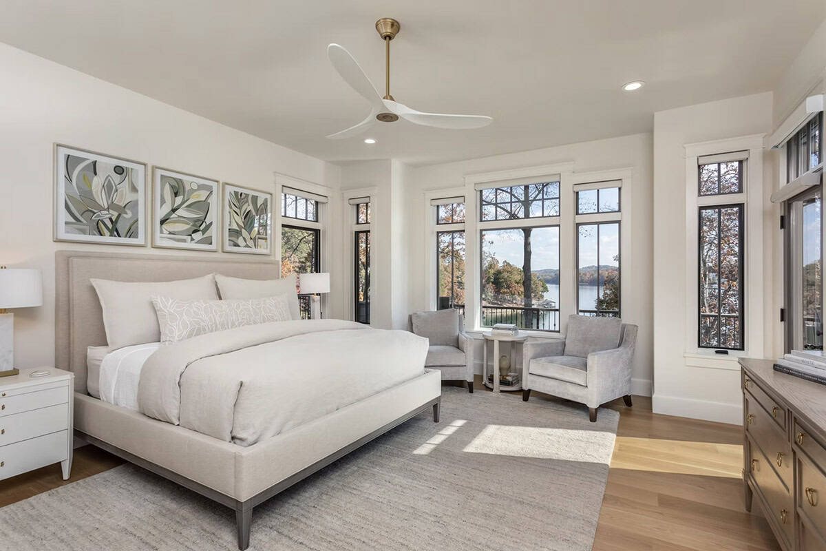 Airy bedroom with lake views, gray armchairs, botanical artwork, and a sleek ceiling fan