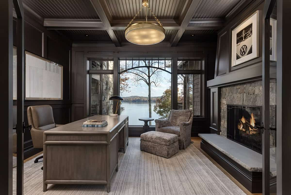 Moody dark-paneled study with lake view, stone fireplace, executive desk, and brass pendant light