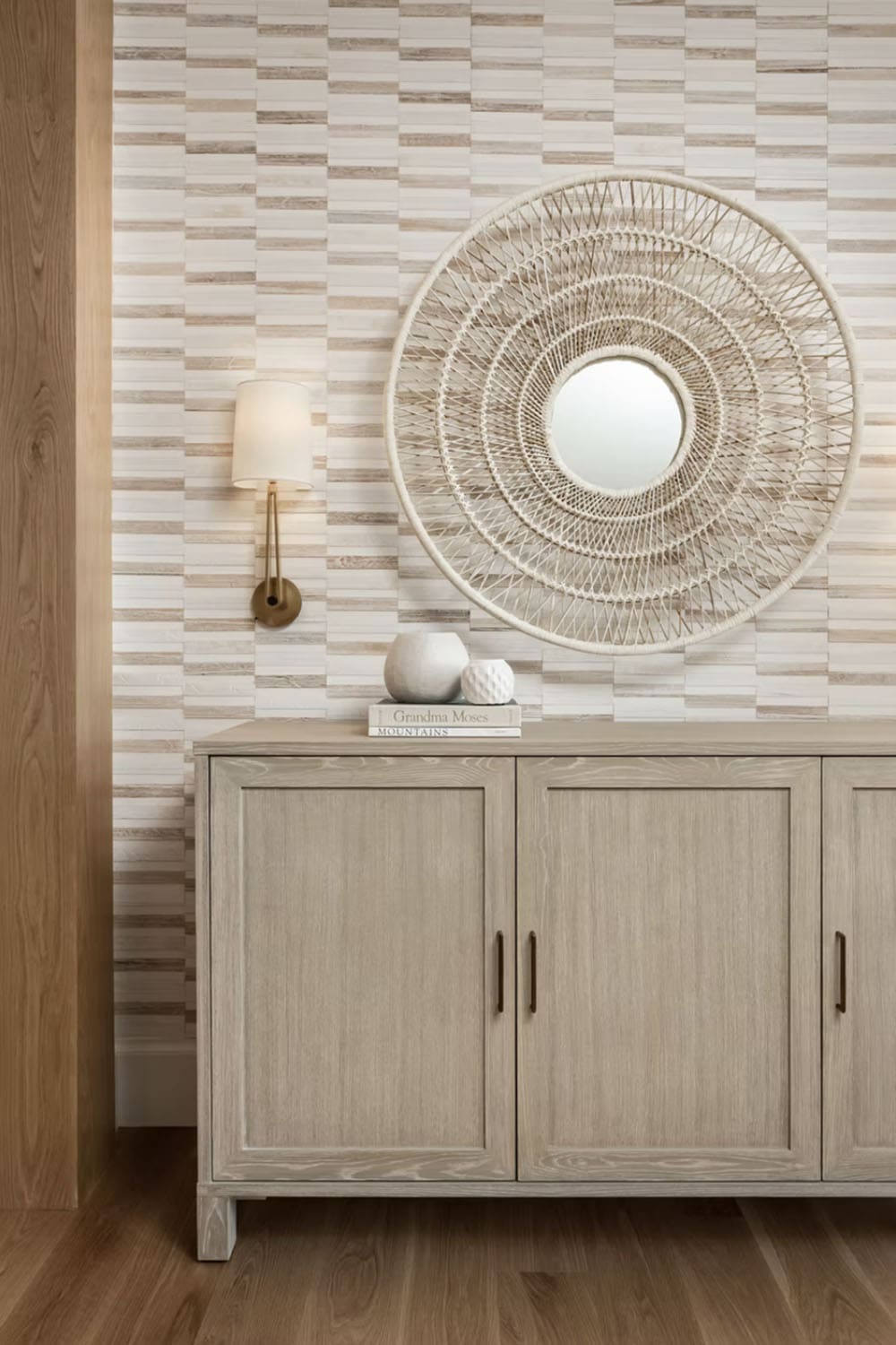 Entryway vignette with whitewashed wood sideboard, woven rattan mirror, and brass wall sconce on a stone mosaic accent wall