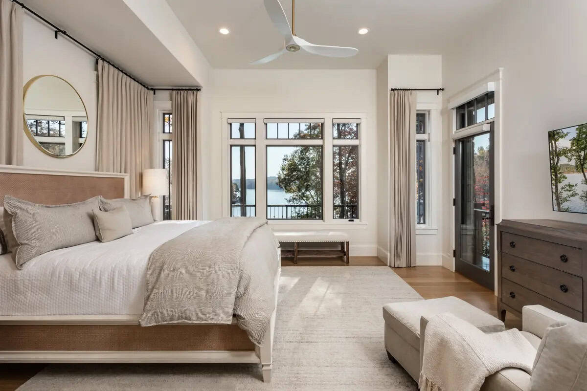 Airy primary bedroom with woven headboard, lake views, round gold mirror, and sleek ceiling fan
