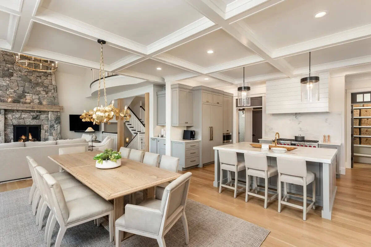 Open-plan dining and kitchen with coffered ceiling, gold leaf chandelier, gray island, and stone fireplace beyond