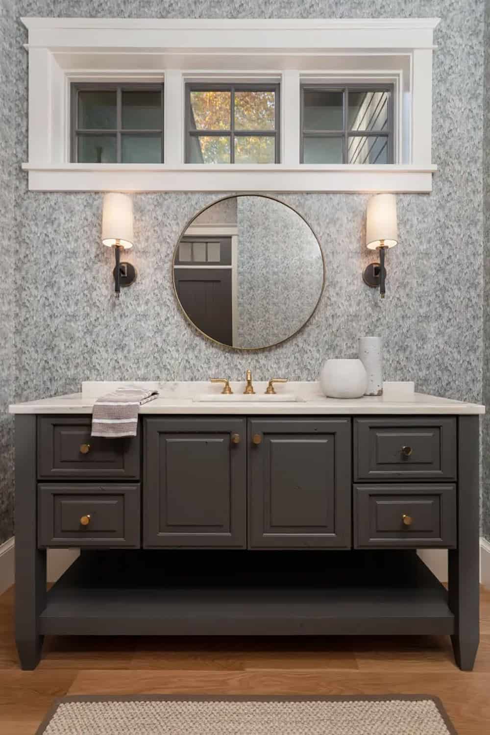 Guest bathroom vanity with dark cabinetry, brass hardware, round mirror, and textured gray wallpaper