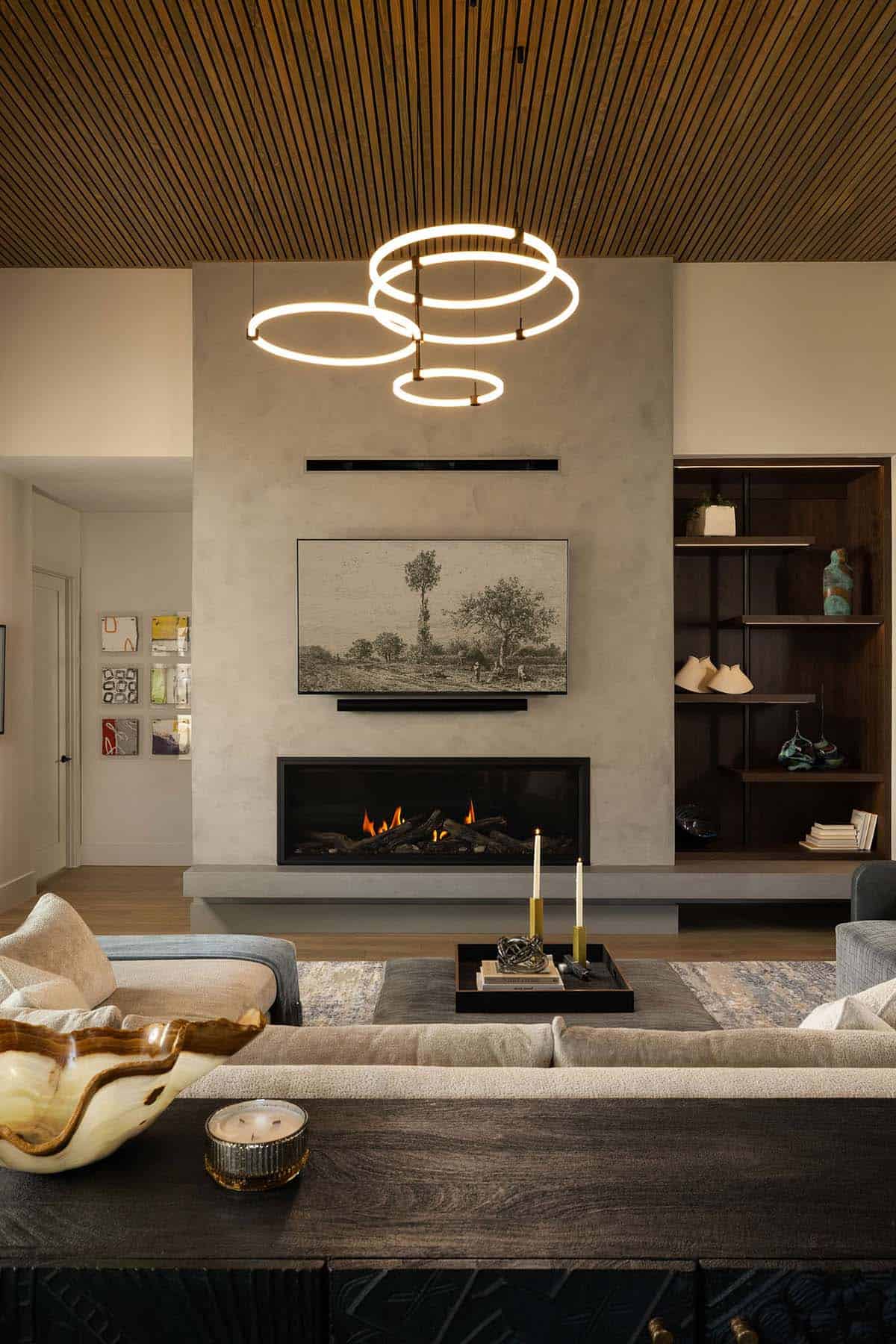 Modern living room with ring LED chandelier, linear gas fireplace, two-tone wood slat ceiling, and dark walnut built-in shelving