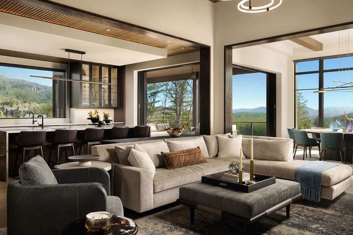 Open plan living room with neutral sectional, mountain views through floor-to-ceiling windows, and dark walnut kitchen beyond