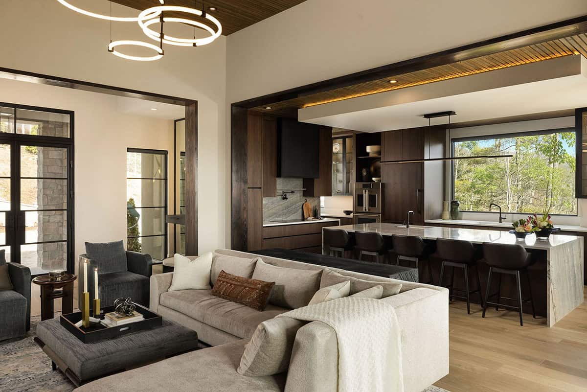 Open plan great room with neutral sectional, ring LED chandelier, dark walnut kitchen island, and black-framed doors to screened porch