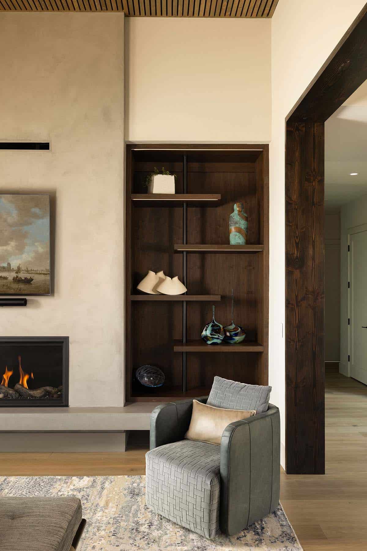 Living room detail showing linear gas fireplace, dark walnut built-in bookcase with art objects, and gray textured accent chair