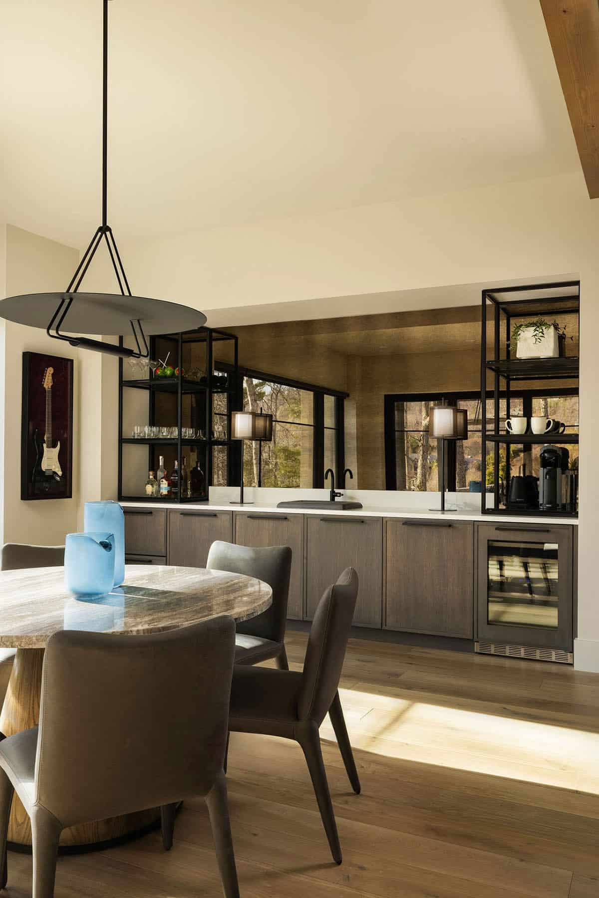 Lower level family room with round stone-top dining table, gray cabinetry wet bar, black steel open shelving, and woodland views