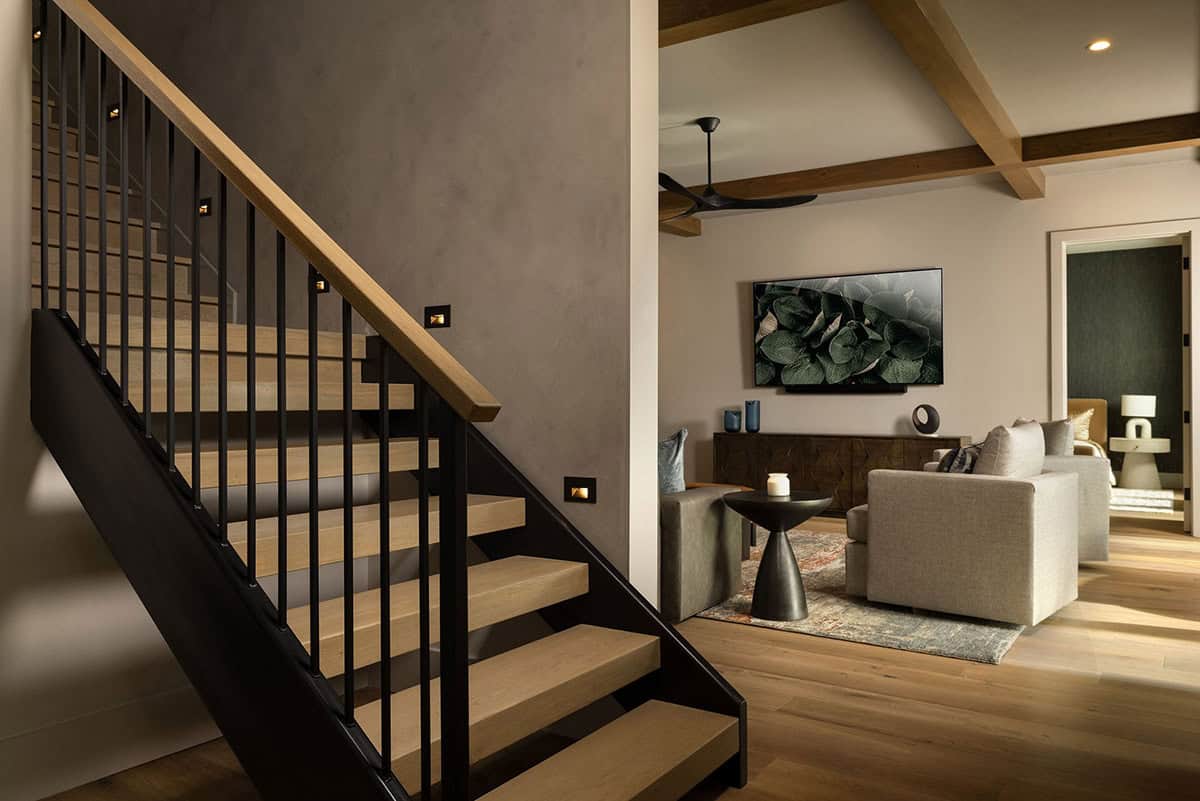 Open oak staircase with black steel balusters and step lights opening to lower level family room with exposed timber beams