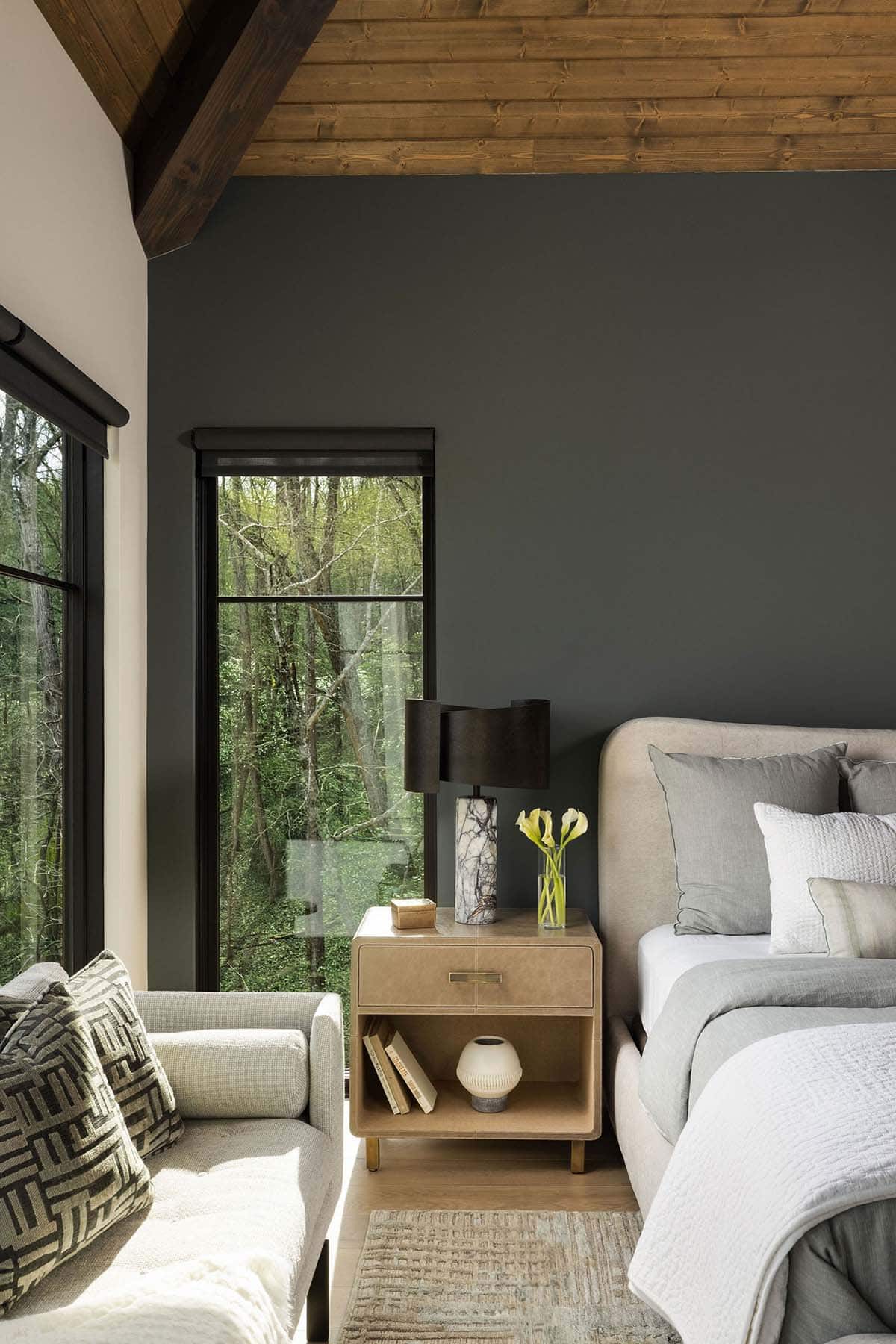 Guest bedroom with dark charcoal accent wall, vaulted wood ceiling, black-framed woodland windows, and neutral upholstered bed