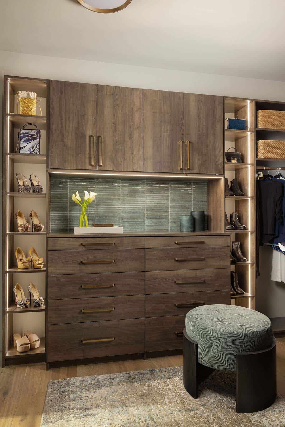 Custom walk-in closet with dark walnut cabinetry, brass hardware, illuminated shoe shelving, green tile backsplash, and velvet ottoman