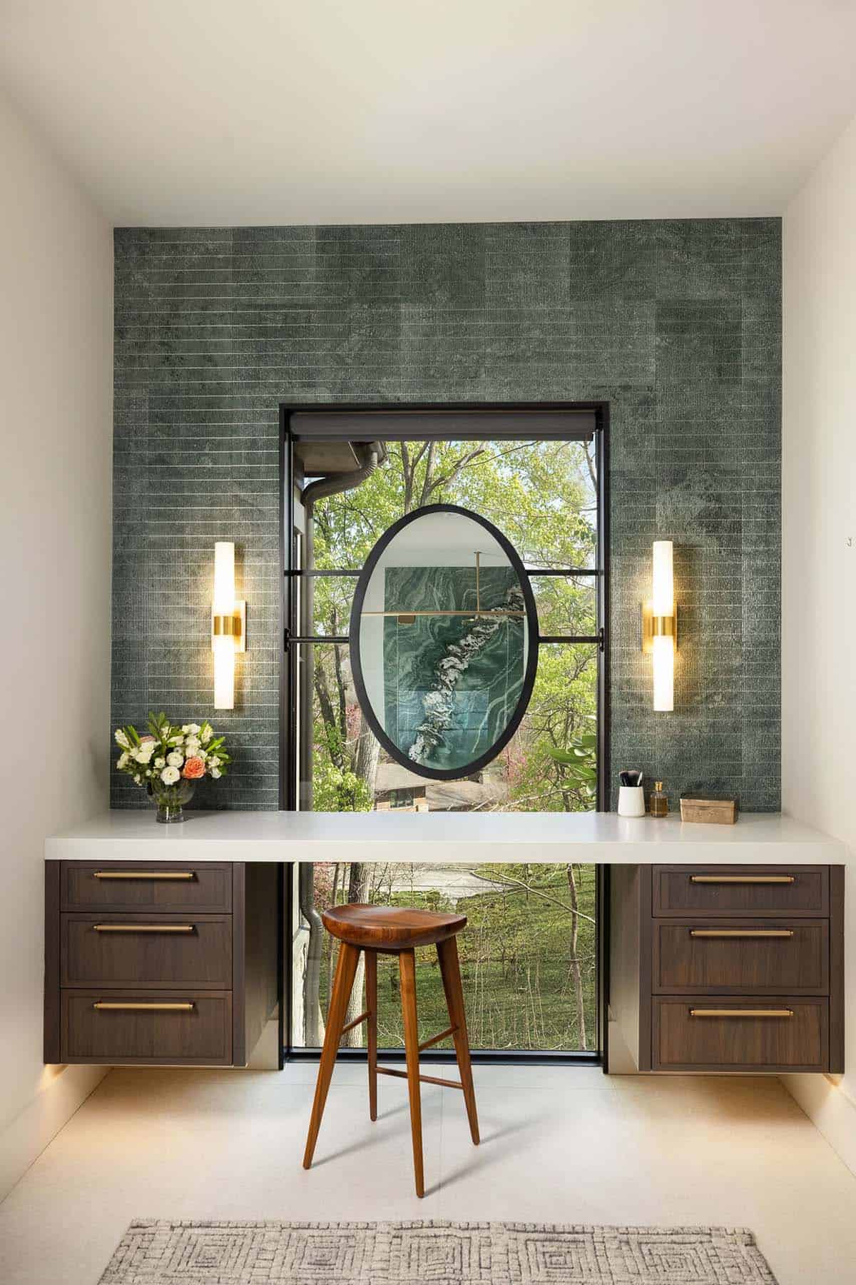 Primary bathroom makeup vanity with green stone tile feature wall, oval mirror, brass sconces, and black-framed woodland window