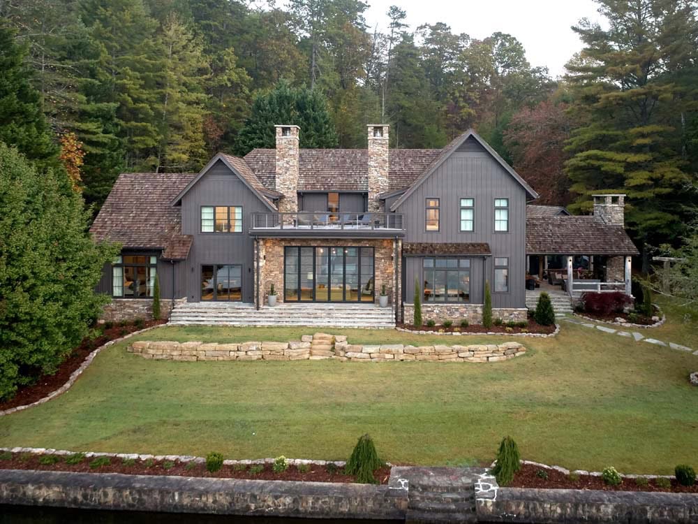 Aerial rear view of gray board-and-batten lake house with stone chimneys and terraced lawn