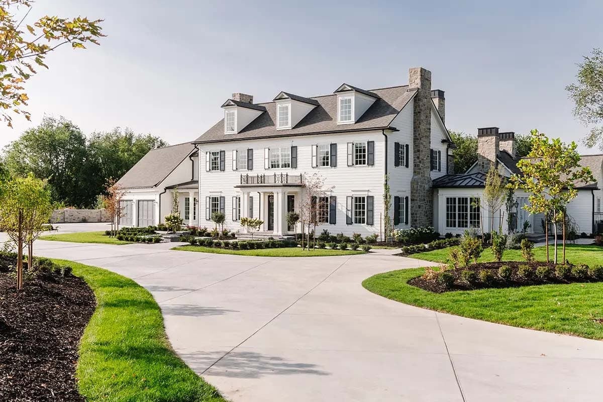 White colonial home with black shutters, dormers, stone chimneys, and circular driveway