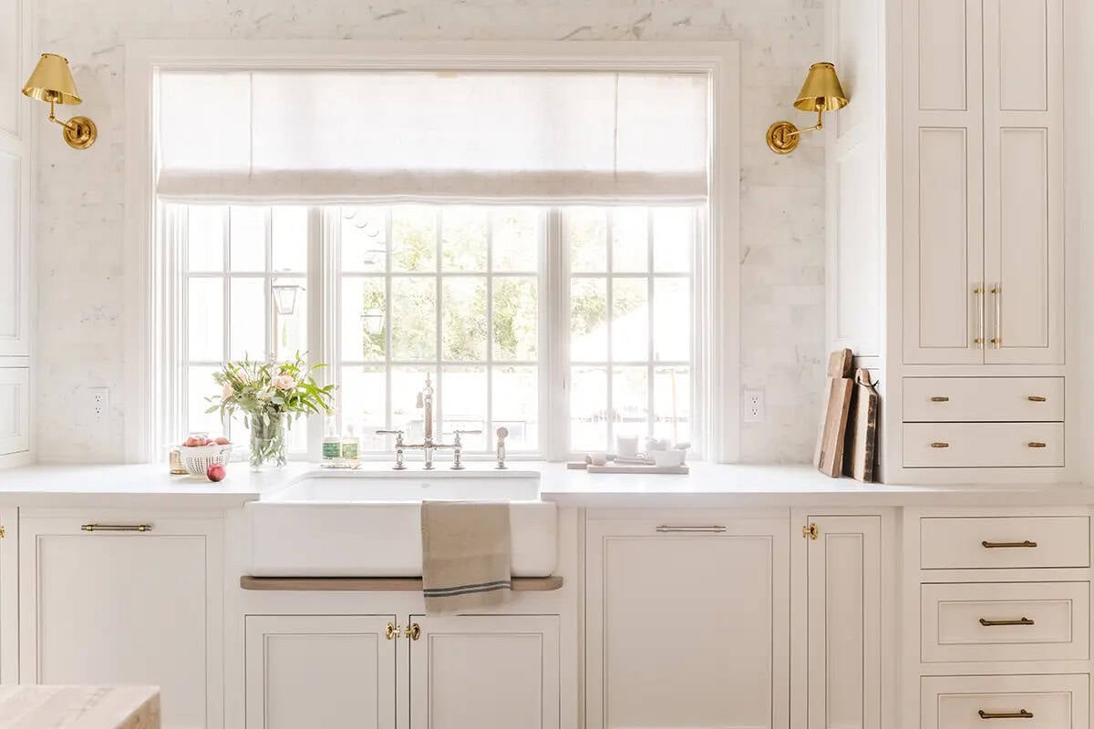 White farmhouse sink with bridge faucet, gold sconces, and marble backsplash