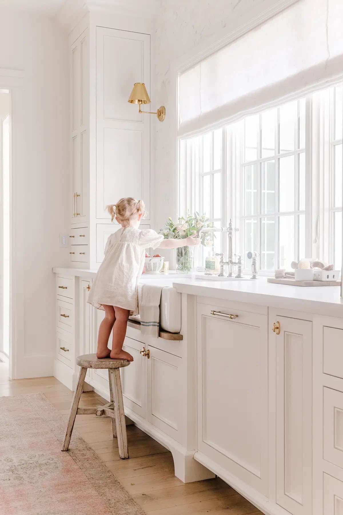 Young girl standing on stool at farmhouse sink in bright white kitchen