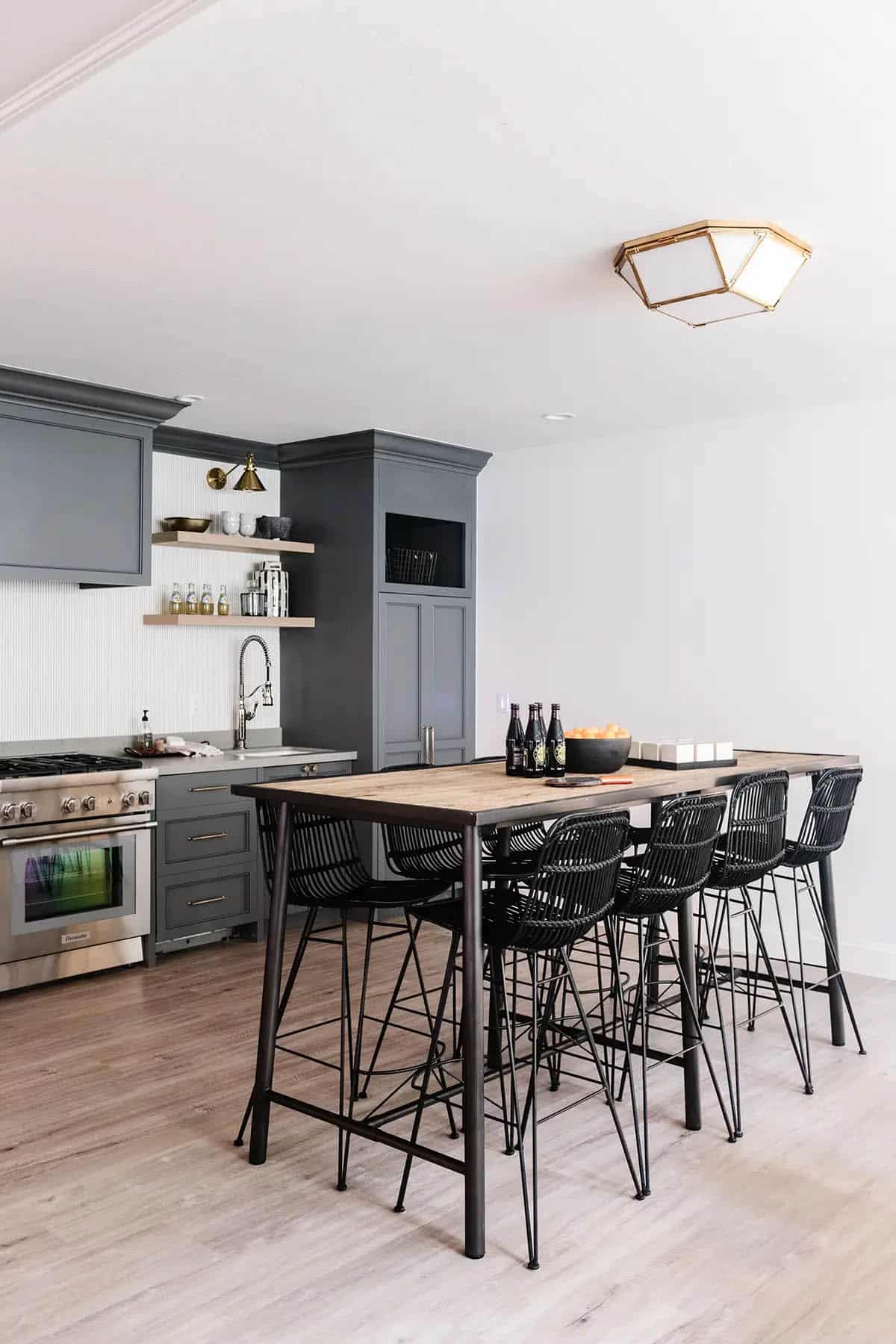 Basement kitchenette with dark gray cabinets, wood bar table, black rattan stools, and stainless range