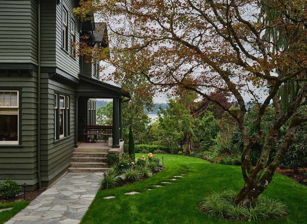 Side view of green craftsman farmhouse with flagstone path, lush garden, and water views through trees