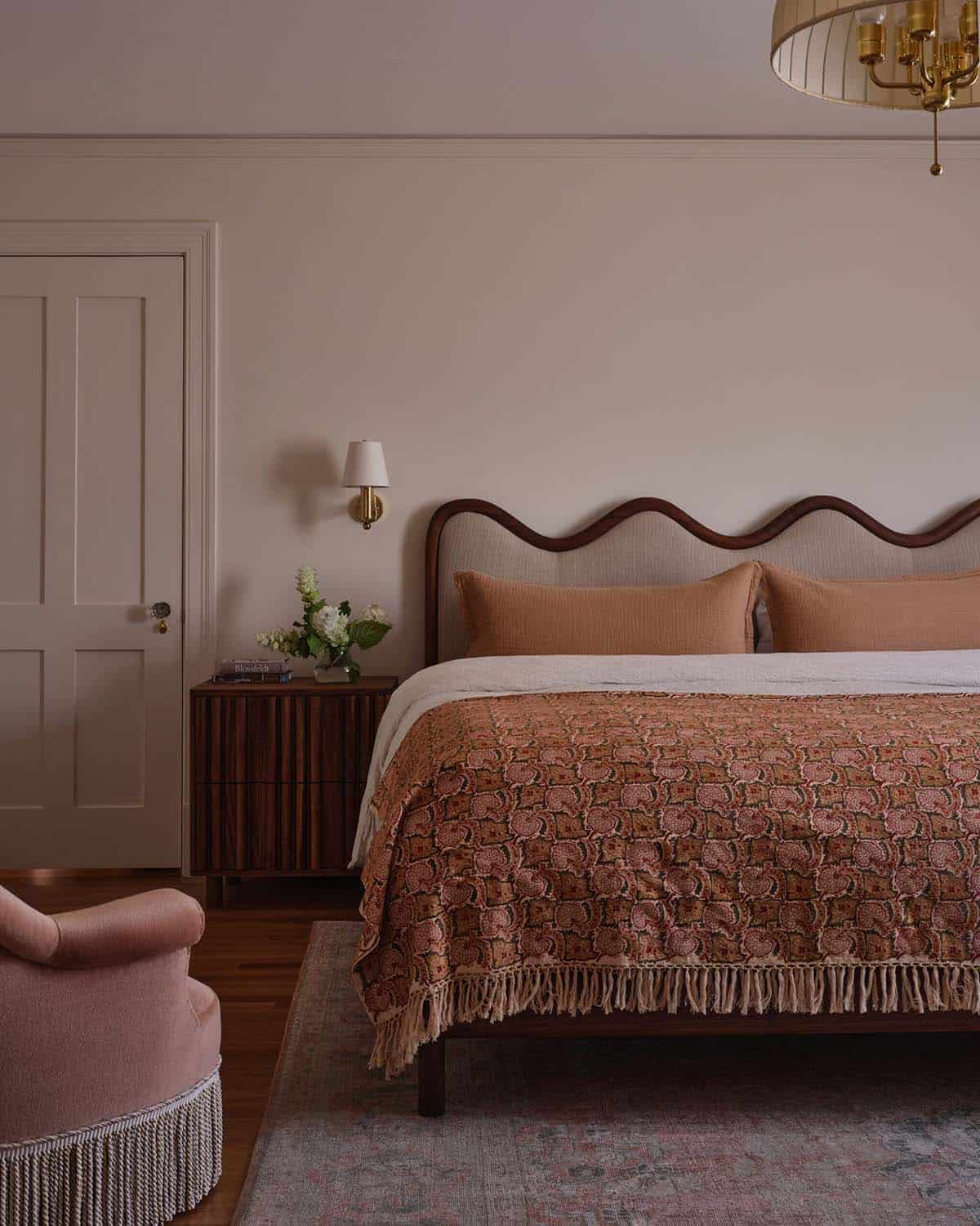 Bedroom with wavy upholstered headboard, terracotta bedding, brass chandelier, and pink velvet chair
