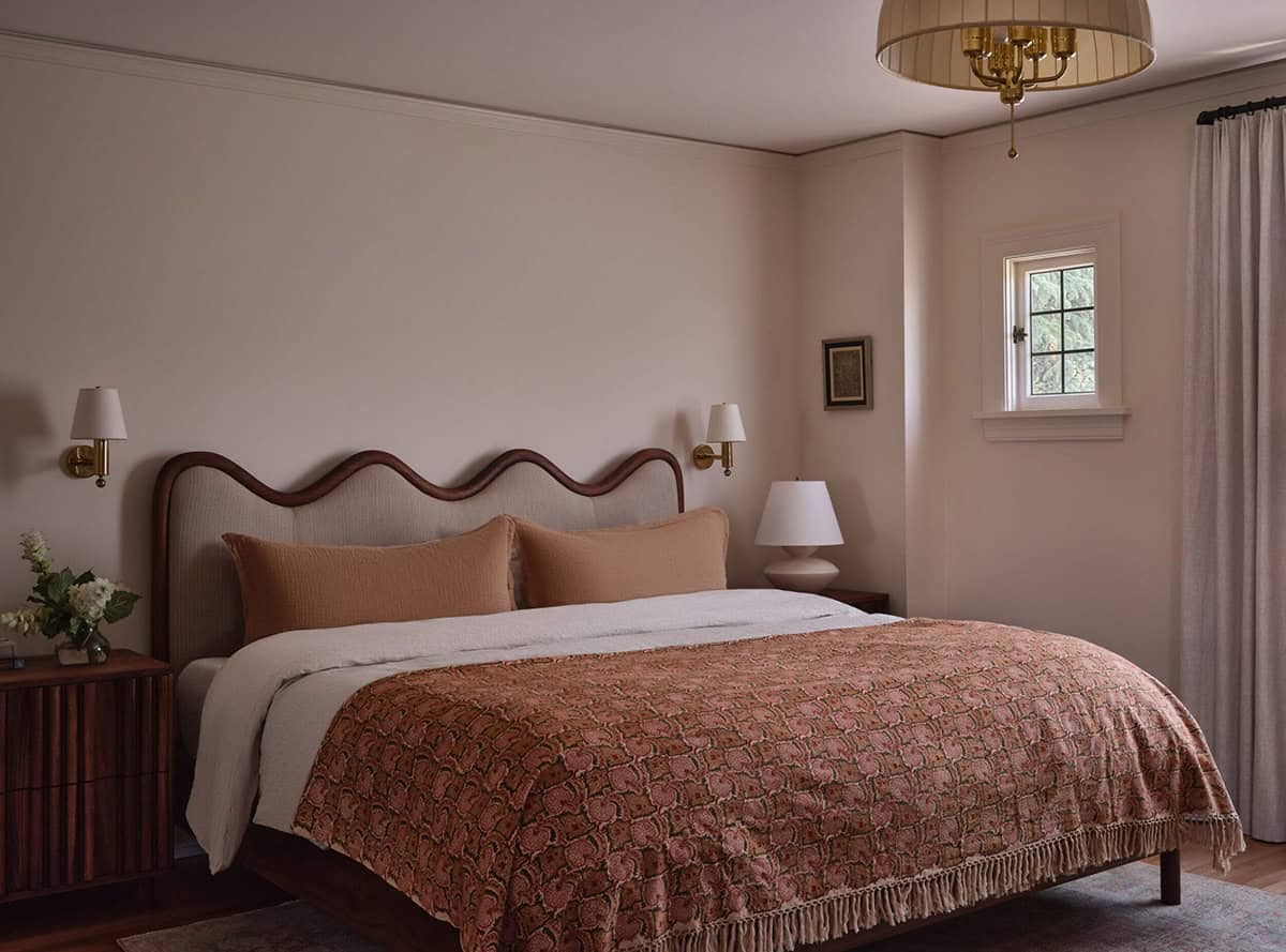 Primary bedroom with wavy upholstered headboard, terracotta quilt, brass sconces, and drum chandelier