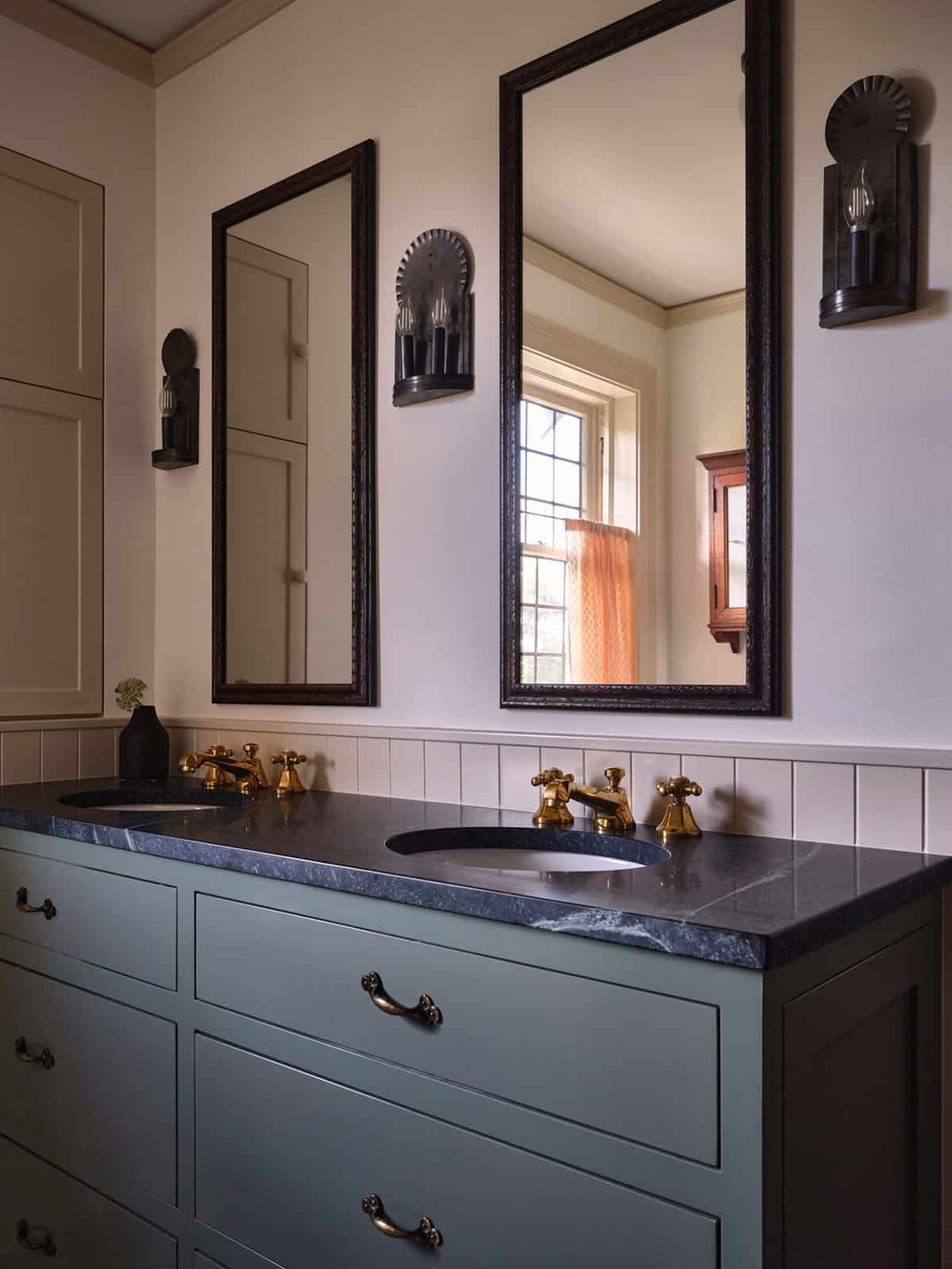 Double vanity with slate-blue cabinets, dark stone countertop, framed mirrors, and brass faucets