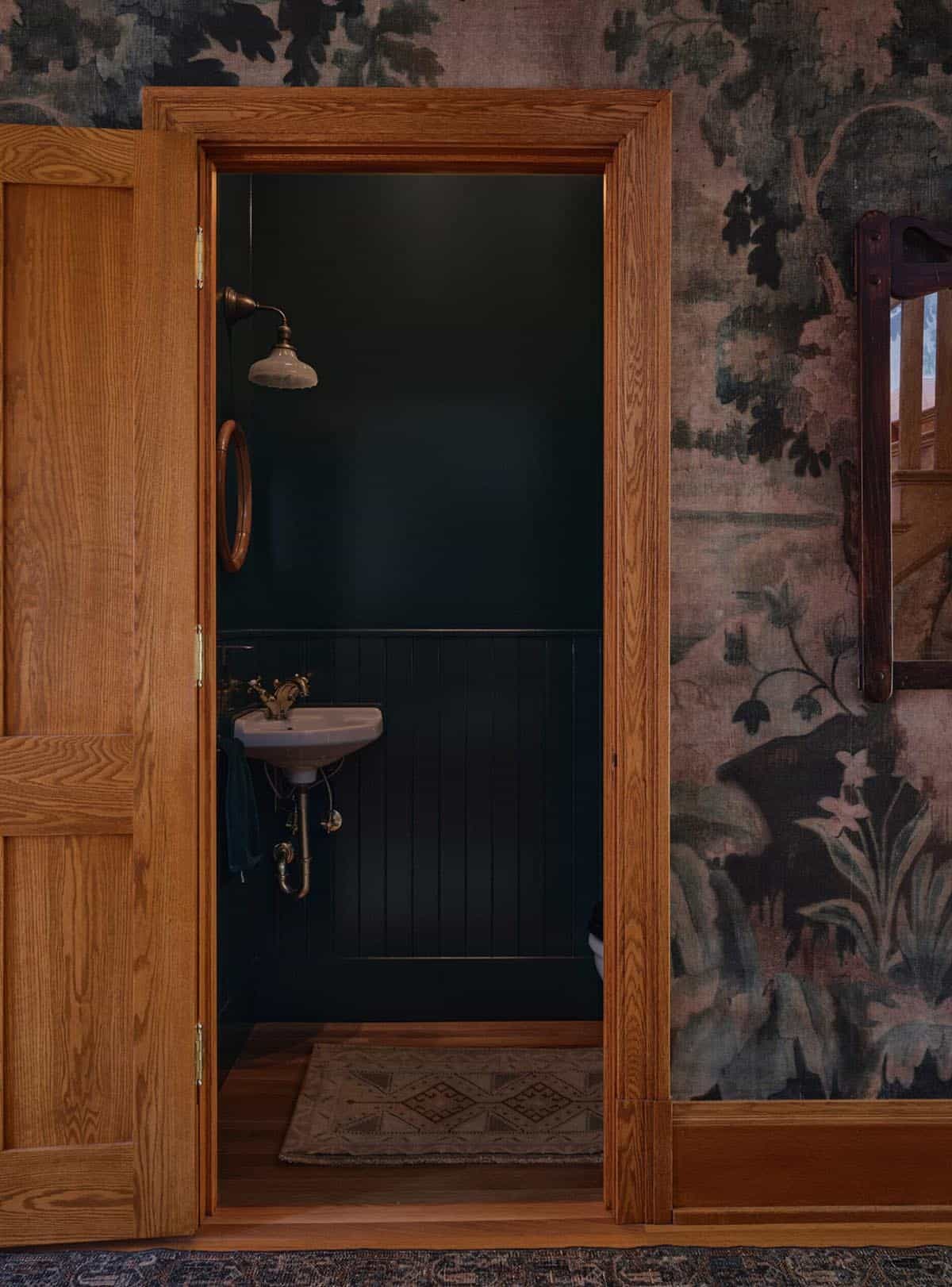 Powder room viewed through oak doorway with dark walls, pedestal sink, and tapestry wallpaper hallway