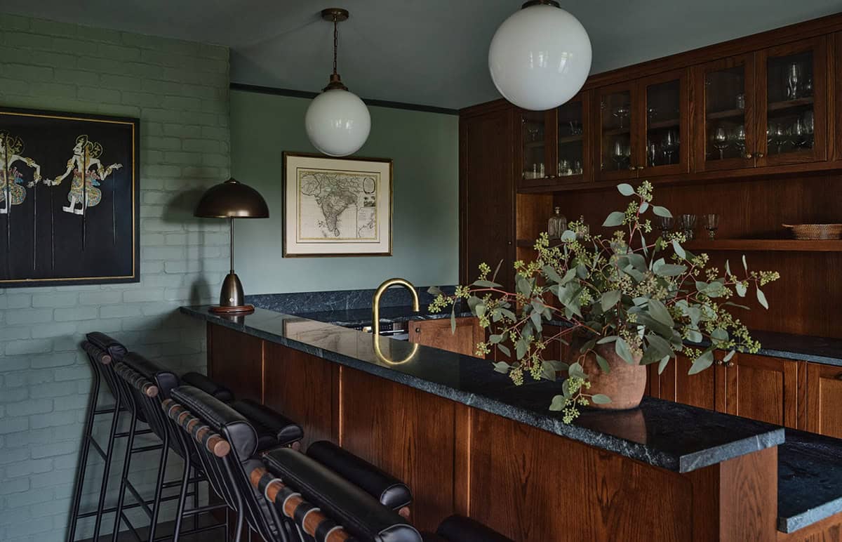 Basement bar with dark soapstone countertop, wood cabinetry, globe pendants, and sage green brick walls