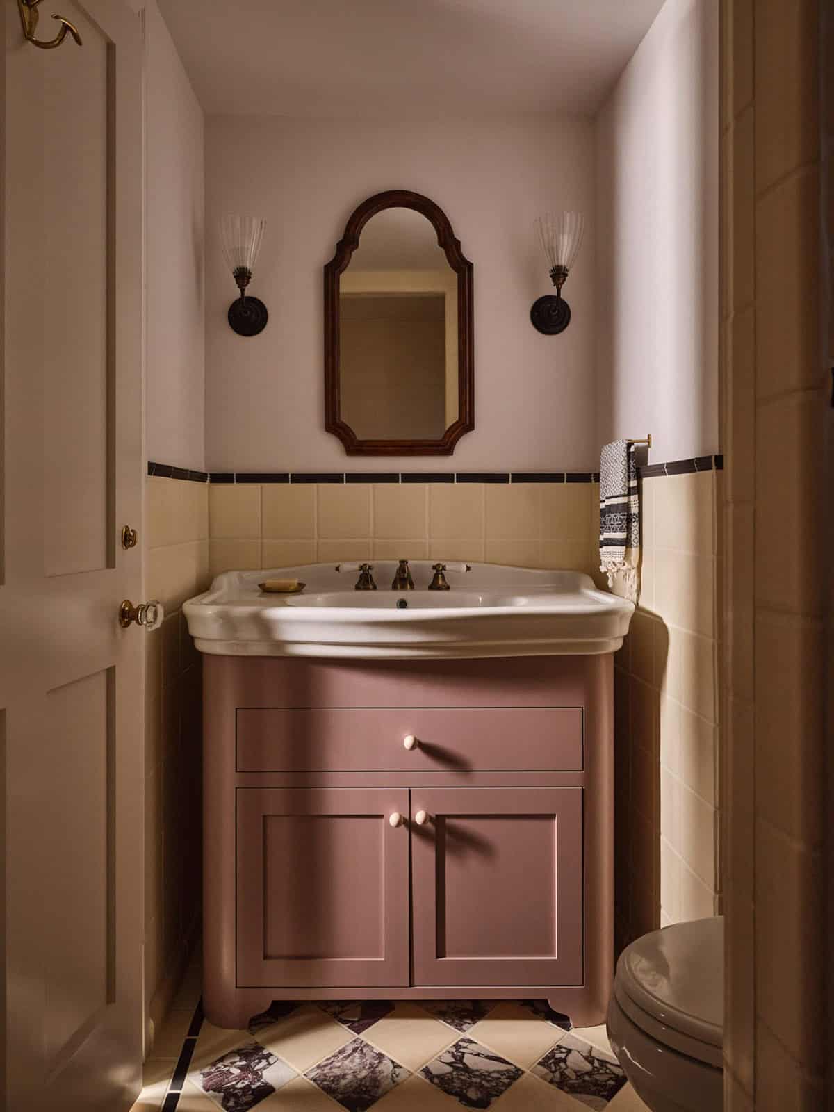 Small bathroom with dusty rose vanity, arched wood mirror, diamond marble tile floor, and sconce lights