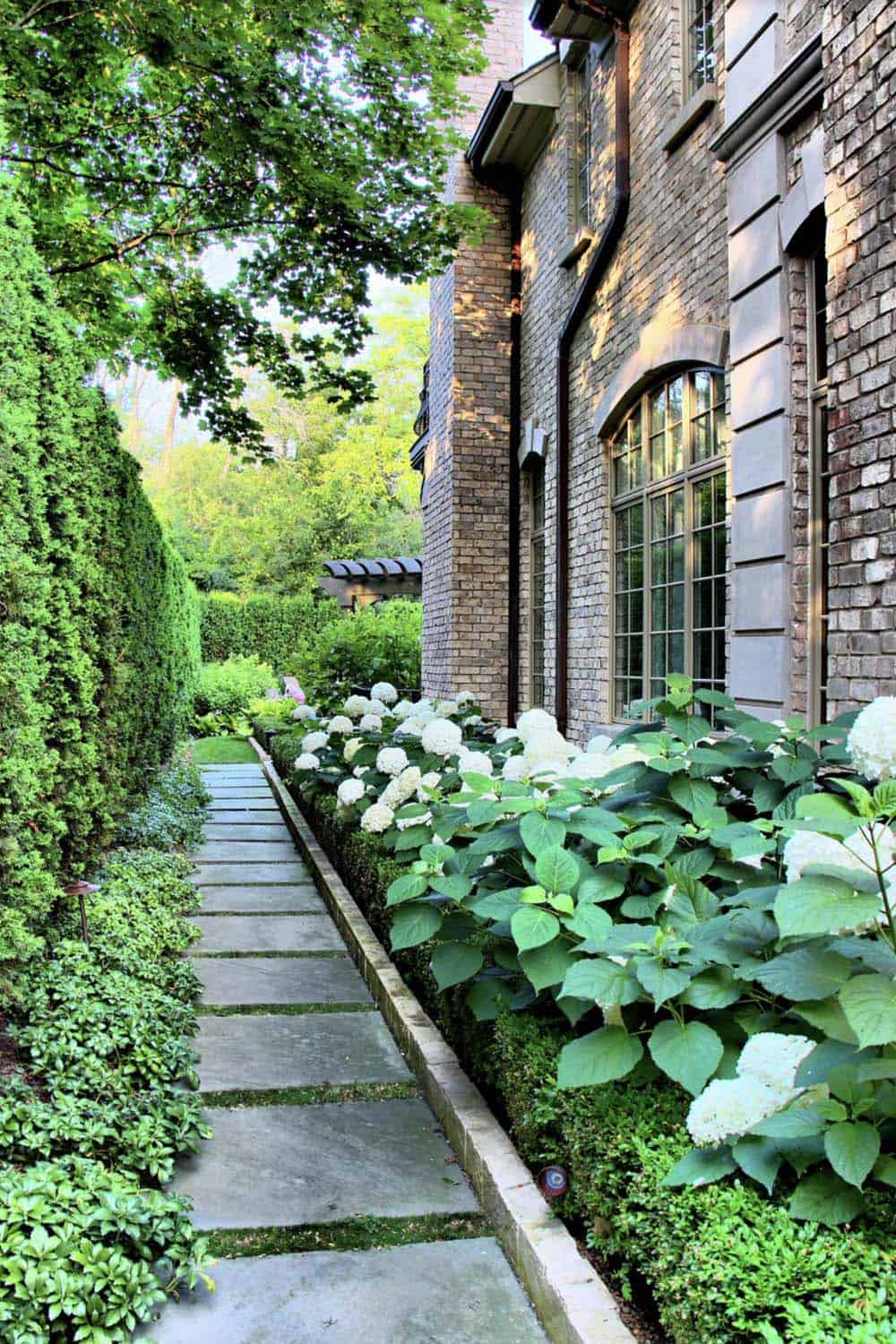 Narrow side garden path with stone pavers flanked by white hydrangeas along a brick house wall and a tall green hedge