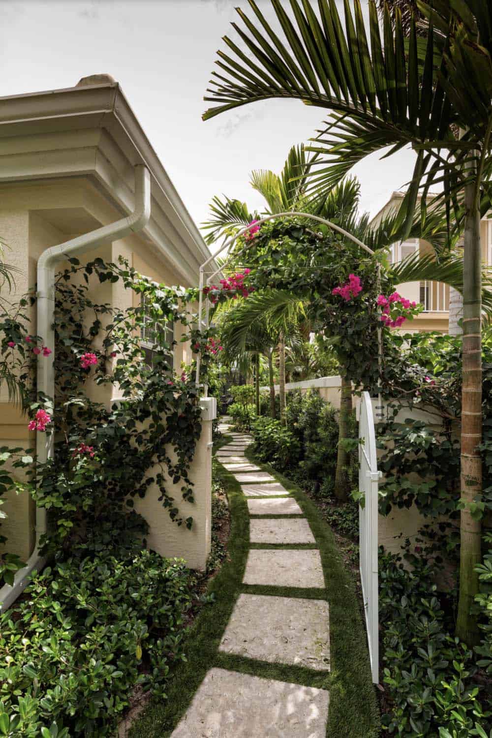 Narrow tropical garden path with stepping stones, bougainvillea climbing over a white arch, and lush greenery between two homes