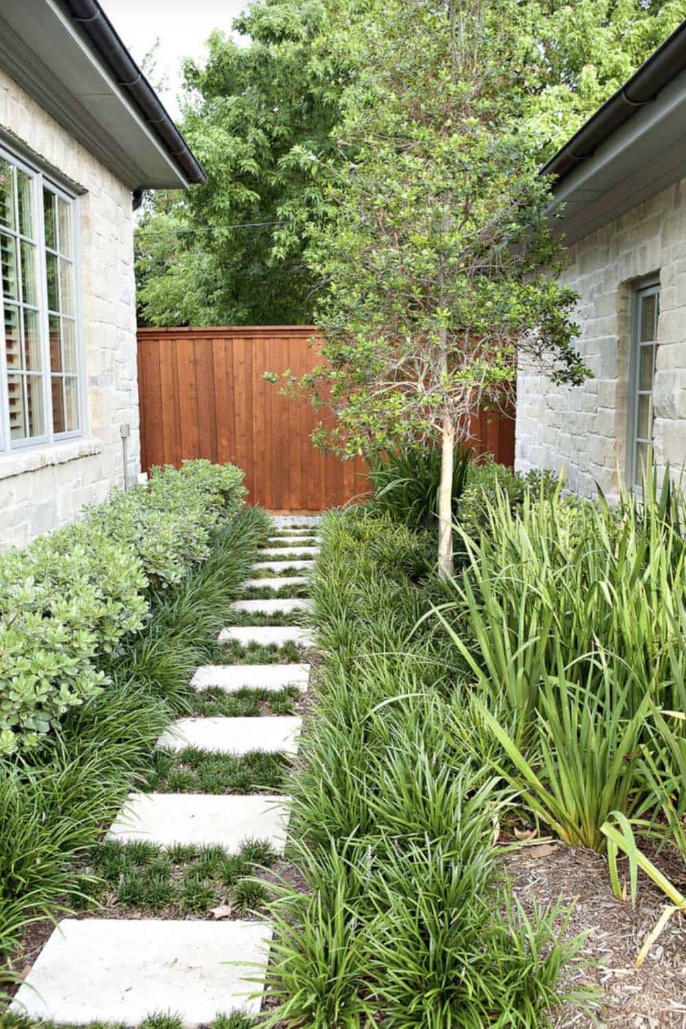 Narrow side yard between two stone homes with stepping stone path, lush ornamental grasses, and a small tree against a wood fence