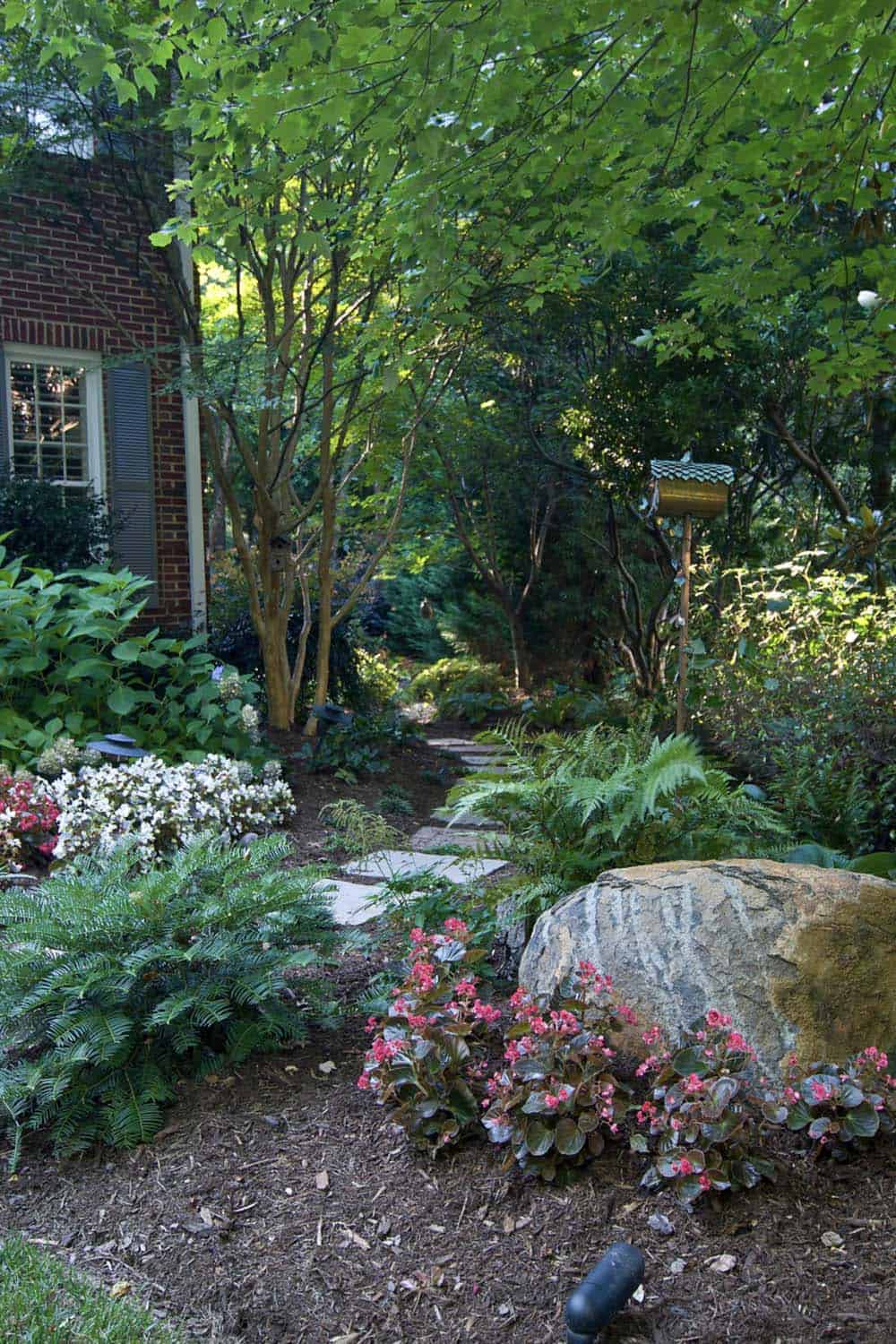 Shaded narrow side yard garden of a brick house in Charlotte with ferns, begonias, azaleas, stepping stones, and boulder accent