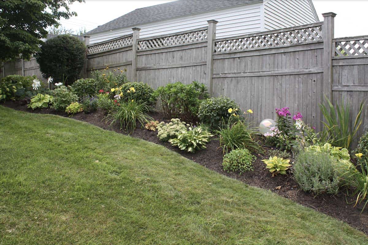 A narrow mixed perennial border along a gray wood privacy fence, featuring hostas, daylilies, and colorful flowering plants against a lush lawn