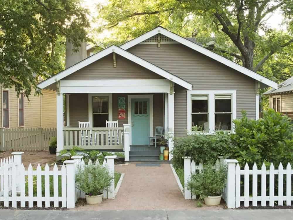 Craftsman bungalow with white picket fence, aqua front door, rocking chairs, and cottage garden plantings