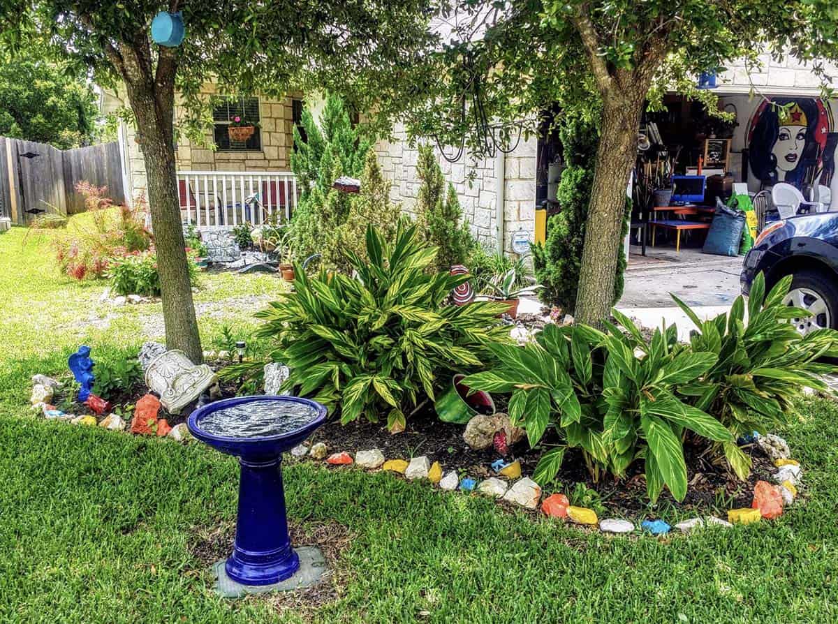 Small front yard with cobalt blue birdbath, colorful rock border, garden sculptures, and tropical plantings