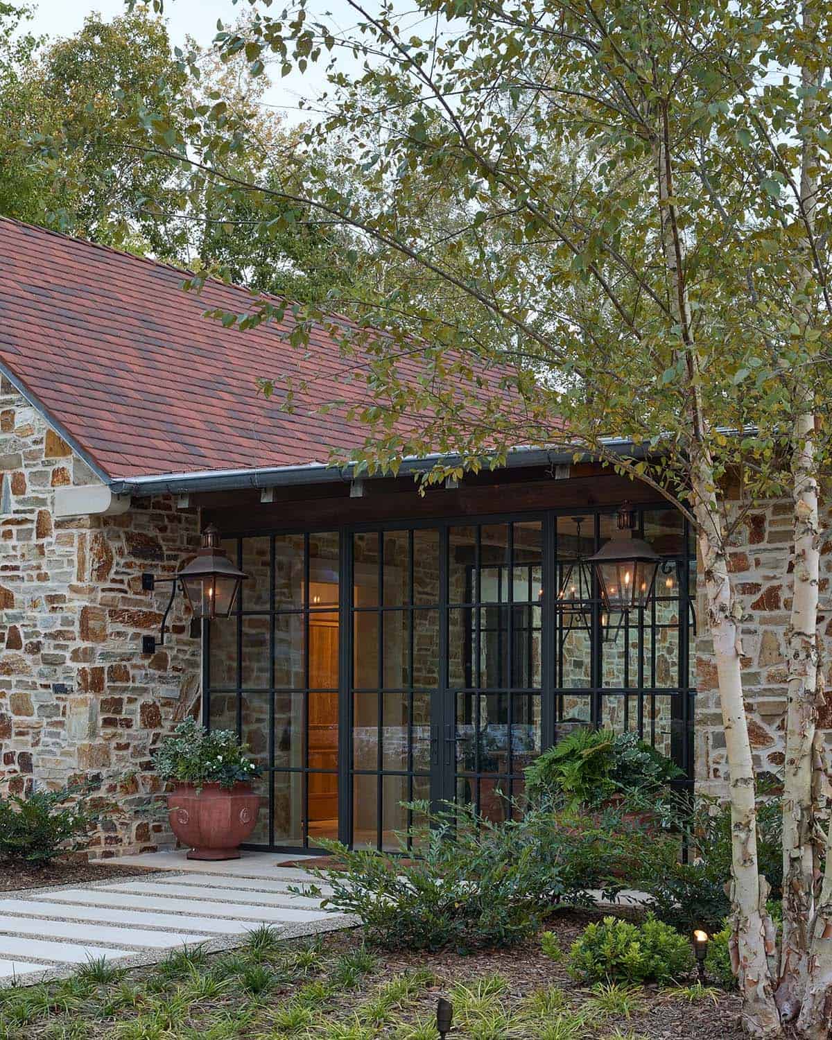 Stone home entrance with steel-frame glazed vestibule, copper lanterns, and birch trees
