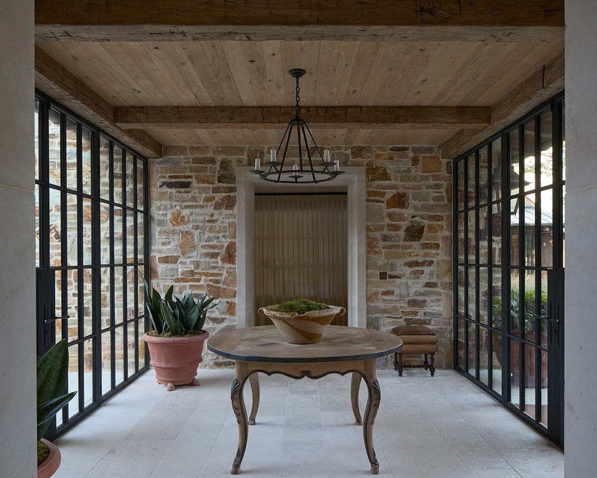 Glass-enclosed foyer with round antique table, iron chandelier, and stone interior walls