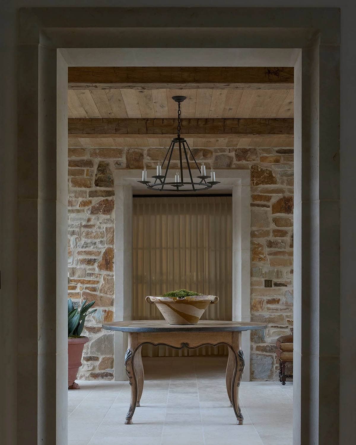 View through doorway into stone foyer with French table, iron chandelier, and pleated curtain beyond