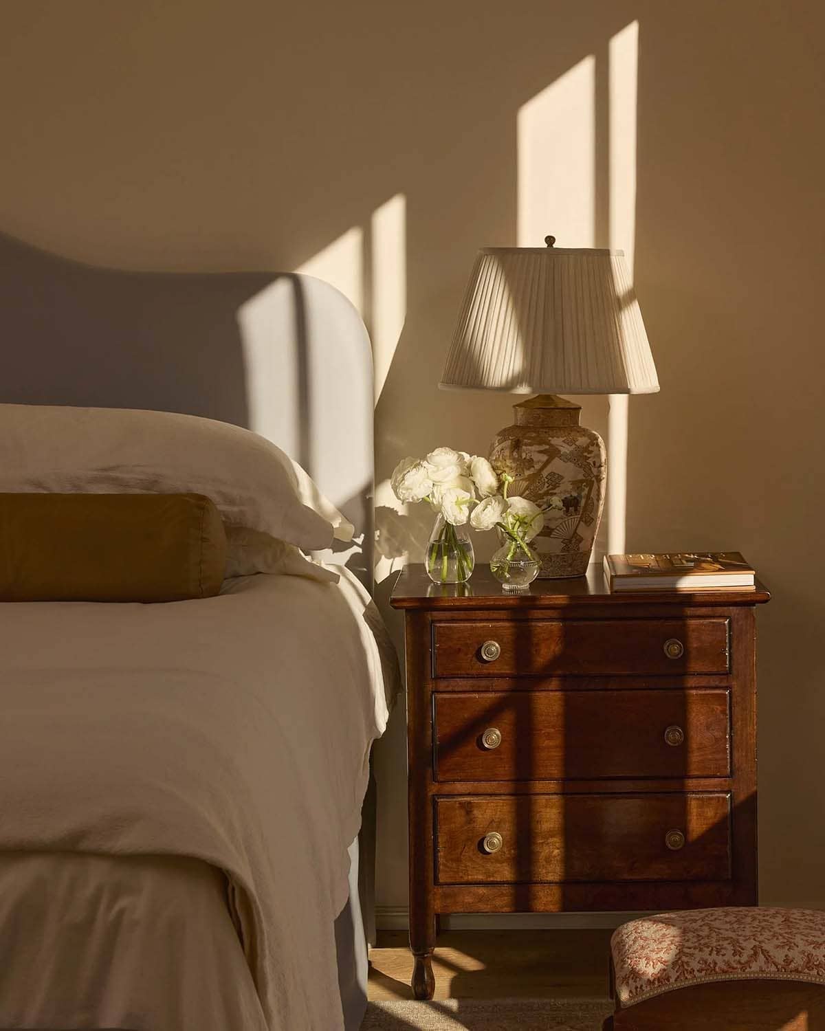 Bedroom nightstand vignette with chinoiserie lamp, white roses, and warm afternoon light on tan walls