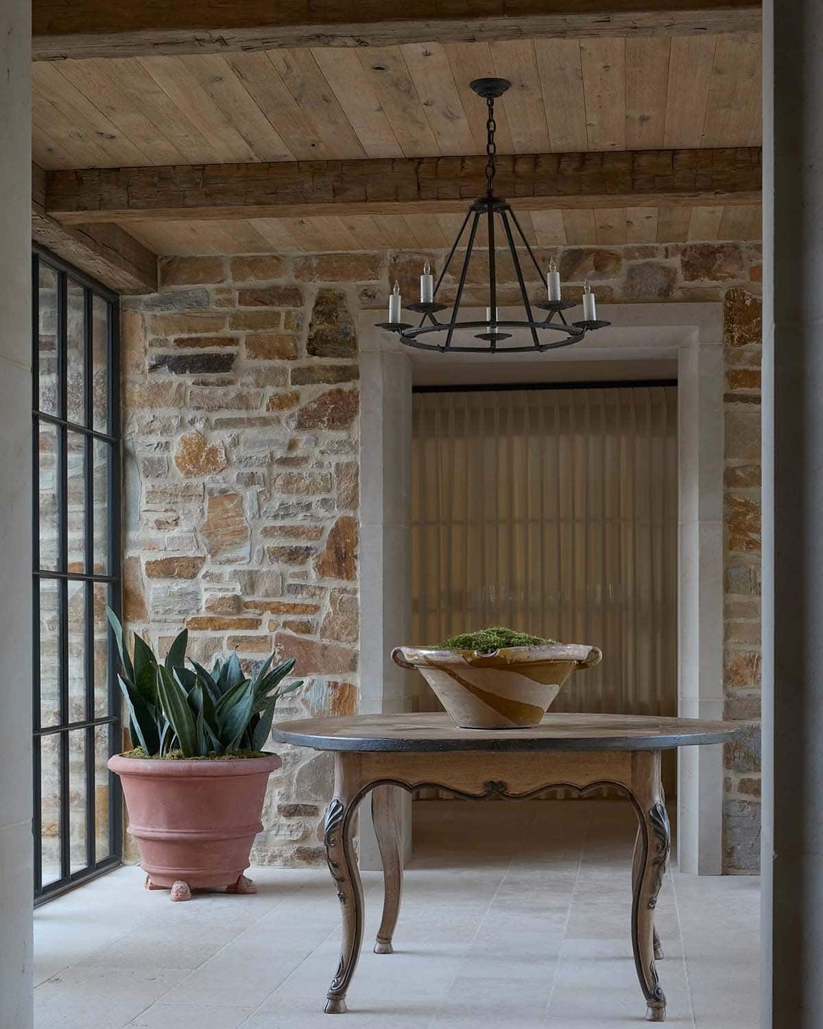 Stone foyer with round French table, terracotta planter, iron chandelier, and steel windows